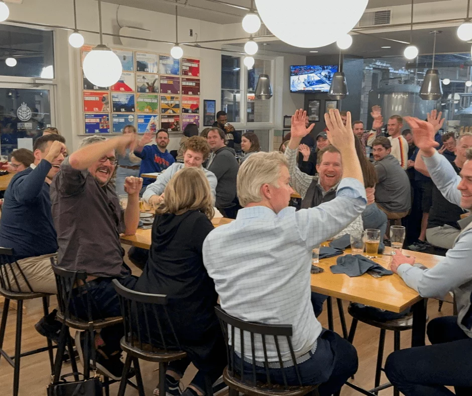 Large group of trivia players raising hands and cheering inside a busy brewery taproom