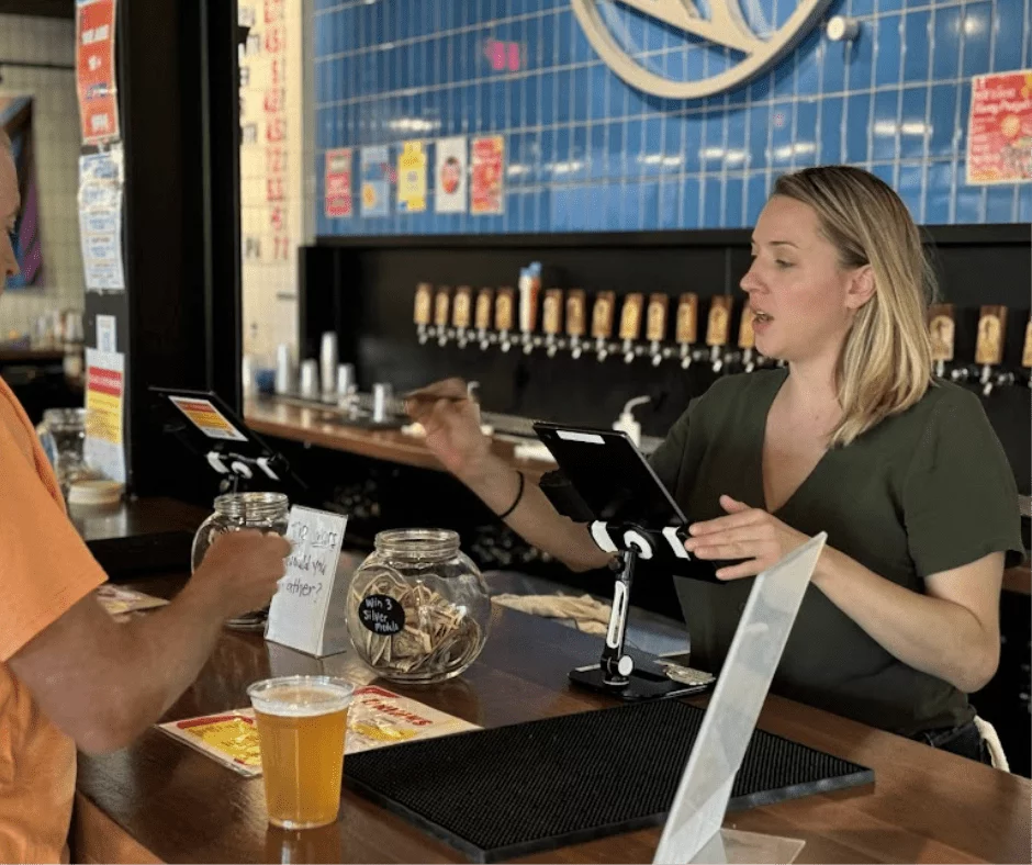 Bartender talking with a guest at a bar counter during a trivia night vote with tip jars and a tablet
